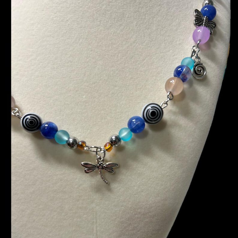 May include: A beaded necklace with a dragonfly pendant. The necklace features a variety of colorful beads, including blue, teal, pink, and black and white spiral beads. A silver dragonfly charm hangs from the center.
