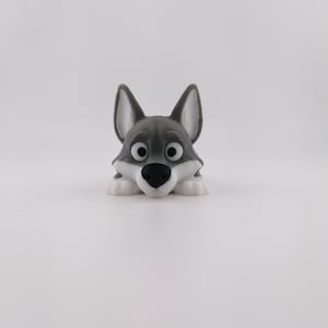 Puede incluir: Figura de lobo gris con pecho y patas blancas, nariz negra y ojos grandes y expresivos. El lobo está tumbado, pareciendo asomarse por el borde. Las orejas son negras con interiores blancos.