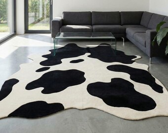 May include: A cream and black cowhide-style rug, featuring large, irregular black patches against a cream backdrop. The rug is laid on a concrete floor in a modern living room, alongside a dark grey sofa and glass coffee table.