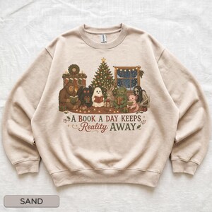 May include: A sand-colored crewneck sweatshirt featuring a graphic illustration of various mythical creatures reading books in front of a Christmas tree. The text reads, A BOOK A DAY KEEPS Reality AWAY, surrounded by small decorative holiday elements.