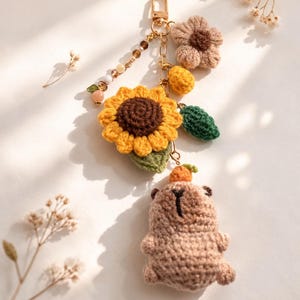 May include: A handmade crochet keychain featuring a sunflower, a bear, and other floral elements. The sunflower has yellow petals and a brown center. The bear is light brown with a small orange carrot on its head. The keychain is attached to a gold-colored clasp.