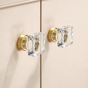 May include: Two cabinet knobs with a clear, square crystal design and gold-colored metal bases. The knobs are mounted on a light beige cabinet door, offering a modern and elegant aesthetic. The hardware adds a touch of luxury.