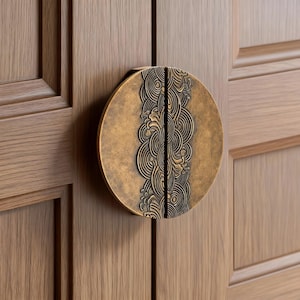 May include: A circular cabinet pull in antique brass, featuring an elaborate, swirling pattern. It's mounted on a light-toned wooden cabinet door. The design is central, offering a decorative and practical hardware solution.