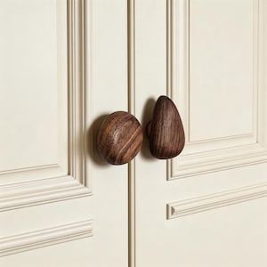 May include: Close-up of two wooden cabinet knobs. One is round, the other is teardrop-shaped. Both are made of dark wood with visible grain, mounted on a cream-colored cabinet door. The knobs are a natural, organic design element.
