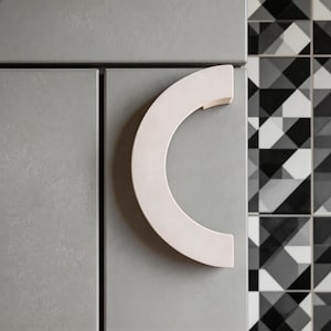 Può includere: Primo piano di un'anta di armadio grigia con una maniglia curva bianco sporco. La maniglia ha la forma di un semicerchio. Lo sfondo presenta un motivo geometrico di piastrelle bianche, nere e grigie.