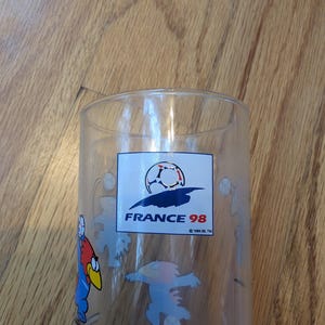 May include: A clear glass tumbler featuring the France 98 soccer logo with a stylized soccer ball graphic and the text FRANCE 98. The glass is decorated with colorful cartoon characters playing soccer, including a red and blue bird-like figure.