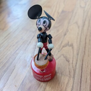 May include: A vintage Mickey Mouse toy drummer. The black and red toy features a Mickey Mouse figure with articulated limbs, holding drumsticks over a small drum. The drum is set on a red base with text around the bottom.