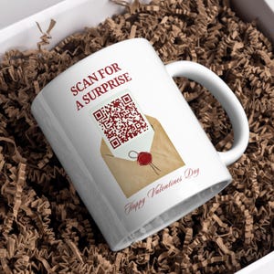 Peut inclure: Mug en céramique blanche avec une anse, portant l'inscription "SCAN FOR A SURPRISE" et "Happy Valentines Day" en bordeaux. Le design comprend un code QR sur une enveloppe avec un sceau de cire rouge.
