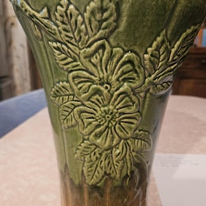 May include: A ceramic vase featuring a glossy olive green upper section with a raised relief pattern of flowers and leaves. The lower portion of the vase transitions into a brown, textured base with vertical fluting. The surface is glazed throughout.