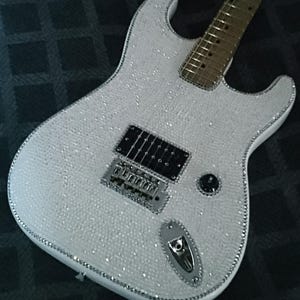 May include: White electric guitar covered in sparkling embellishments. The guitar features a silver trim, black pickups, and a silver bridge. The body is covered in small, reflective crystals, giving it a glamorous appearance. The fretboard is a light brown color.