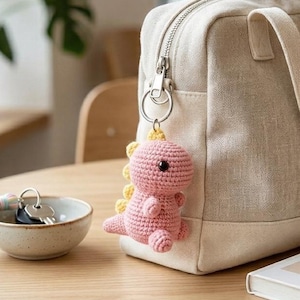 May include: A beige tote bag with a zip and a small pink crocheted dinosaur keyring. A small bowl with keys and a tassel, and a book with a botanical illustration are on a wooden table.