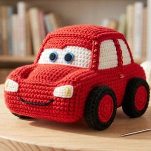 Könnte beinhalten: Ein handgefertigtes rotes Häkelauto mit weißen Akzenten, schwarzen Rädern und blauen Augen. Das Spielzeugauto steht auf einem Holztisch. Rote und weiße Wollknäuel befinden sich im Hintergrund.