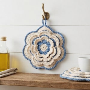 May include: A decorative, flower-shaped crochet potholder in cream and blue hangs on a wall hook. A matching coaster set and a white mug sit on a wooden shelf. A bottle of oil and a small bowl are also on the shelf.