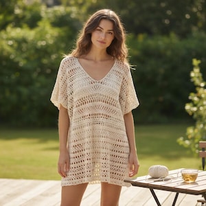 Puede incluir: Una túnica de crochet color crema con tejido abierto, cuello en V y mangas cortas. La túnica llega a la rodilla y tiene un corte holgado y fluido. La imagen está tomada al aire libre, con una mesa de madera y vegetación de fondo.