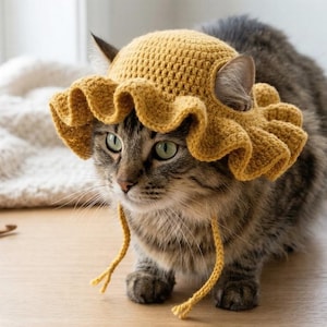 Pode incluir: Um gato malhado usando um chapéu de crochê amarelo mostarda com uma aba franzida e laços de queixo. O gato está sentado em uma superfície de madeira, com fios e suprimentos de crochê ao fundo. O chapéu é o ponto focal.