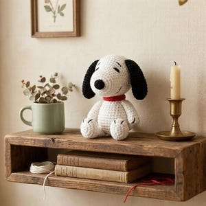 May include: A white and black crocheted Snoopy dog with a red collar sits on a rustic wooden shelf. The shelf also holds books, a candle in a brass holder, and a green mug. A framed botanical print hangs above.