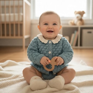 May include: A smiling baby wearing a light blue knitted romper with a cream-colored collar and matching socks. The baby is holding a wooden teething ring and sitting on a cream-colored blanket. A crib and a teddy bear are in the background.