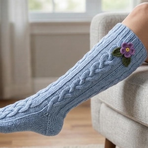 May include: Light blue cable-knit leg warmers with a decorative purple flower and green leaves. The leg warmers are knee-high and feature a textured cable design. The person is sitting in a gray armchair.