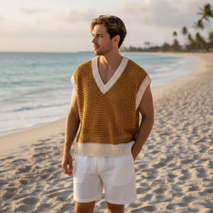 May include: A mustard yellow, open-weave vest with a cream-coloured trim and a V-neck. The vest is paired with white shorts. The person is standing on a sandy beach with the ocean in the background.