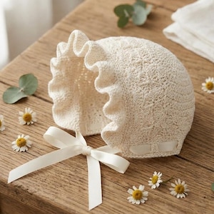 Könnte beinhalten: Eine cremefarbene, gehäkelte Babyhaube mit Rüschenrand und Satinbandschleife. Die Haube liegt auf einer rustikalen Holzoberfläche, verziert mit kleinen Gänseblümchen und Eukalyptusblättern.