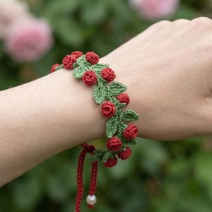 Puede incluir: Una pulsera de ganchillo con capullos de rosa rojos y hojas verdes. La pulsera está atada con un cordón rojo y una perla blanca. La pulsera se lleva en la muñeca.