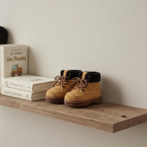 May include: A pair of handmade crochet baby boots in a mustard yellow color with brown and black accents. The boots have laces and a rolled-down cuff. They are displayed on a wooden shelf with books in the background.