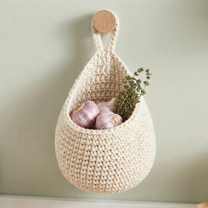 May include: A cream-coloured, crocheted hanging basket filled with garlic bulbs and sprigs of herbs. The basket is suspended from a wooden peg on a light green wall, offering a rustic, handmade aesthetic for kitchen storage.