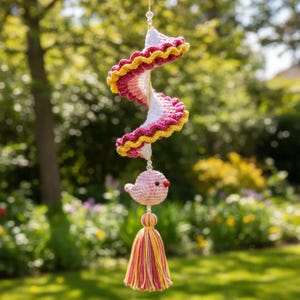 Puede incluir: Móvil de ganchillo hecho a mano con un diseño en espiral en blanco, rosa y amarillo. Un pequeño pájaro rosa con ojos negros y pico rojo cuelga debajo, junto con una borla de hilo amarillo, rosa y naranja. El móvil está suspendido por un hilo transparente.