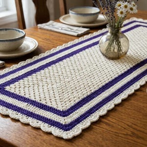May include: A cream-colored, rectangular crochet table runner with a purple border and scalloped edges. The runner is placed on a wooden table, alongside tableware and a vase of flowers. The table setting is for a dining room.