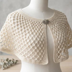 以下が含まれることがあります： クリーム色の、ダイヤモンド模様のデザインのオープンフロントかぎ針編みケープレット。ケープレットは装飾的なシルバー色のブローチで留められています。マネキンに着せられています。