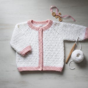 May include: A white and pink crochet baby cardigan with a button-up front. The sweater has a round neckline and cuffs in a soft pink color. A ball of white yarn, a crochet hook, and wooden buttons are also visible.