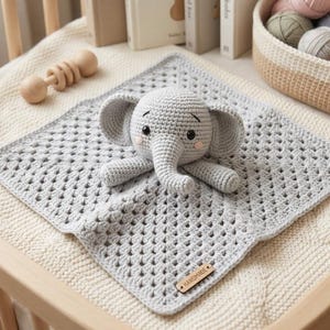 Puede incluir: Una manta de bebé de ganchillo hecha a mano con un juguete de elefante gris. La manta es cuadrada con un patrón de encaje. El elefante es gris con ojos negros y mejillas rosadas. También se ven una sonaja de madera y una cesta de hilo.