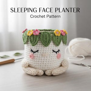 Puede incluir: Una maceta de crochet blanca con una cara durmiente, ojos cerrados, mejillas rosadas y una nariz pequeña. La maceta está adornada con hojas verdes y flores rosas y amarillas alrededor del borde. El texto "SLEEPING FACE PLANTER Crochet Pattern" está en la parte superior.