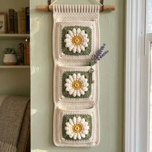 Może przedstawiać: Kremowy, szydełkowy organizer ścienny z trzema kieszeniami, z których każda ma wzór stokrotki z białymi płatkami i żółtym środkiem. Organizer wisi na drewnianym kołku i ma gałązkę lawendy i klucz.