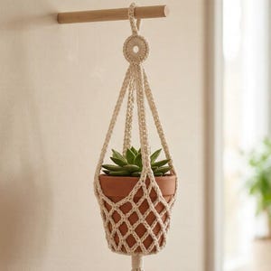 Può includere: Un portavaso in macramè color crema con un vaso in terracotta contenente una succulenta verde. Il portavaso è sospeso a un tassello di legno fissato a una parete beige. Il macramè presenta un motivo a rombi.