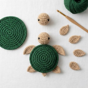 Puede incluir: Una tortuga de ganchillo en varias etapas de montaje. El cuerpo de la tortuga es verde oscuro, con aletas y cabeza beige. Incluye un círculo verde, hilo y un ganchillo. La tortuga está hecha de hilo.