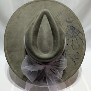 May include: Olive green fedora hat with a wide brim, featuring a moth and moon design. The hat has a white ribbon and a sheer, light pink bow. The hat is a stylish accessory.