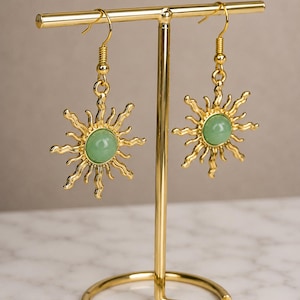 May include: Gold-colored sunburst earrings with a central, round, light green stone. The earrings are displayed on a gold-colored earring stand. The sunburst design features pointed rays extending outwards from the central stone.