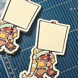 May include: Sticker of a cartoon character holding a blank sign. The character is dressed in a yellow shirt, brown trousers, and a striped hat. The sticker has a white border and is on a blue cutting mat.