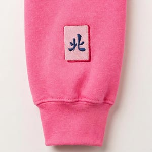 May include: Close-up of a bright pink sweatshirt sleeve. A small rectangular patch with a blue embroidered symbol is sewn onto the sleeve. The cuff is a darker shade of pink.