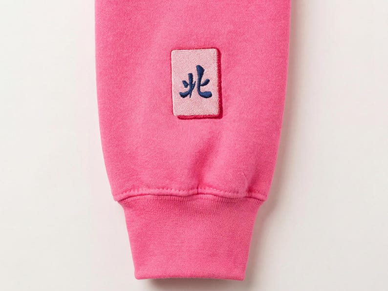May include: Close-up of a bright pink sweatshirt sleeve. A small rectangular patch with a blue embroidered symbol is sewn onto the sleeve. The cuff is a darker shade of pink.