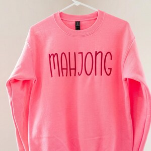 May include: A vibrant pink sweatshirt featuring the word "MAHJONG" in a darker pink, cursive script. The garment is suspended on a white hanger, set against a simple backdrop.