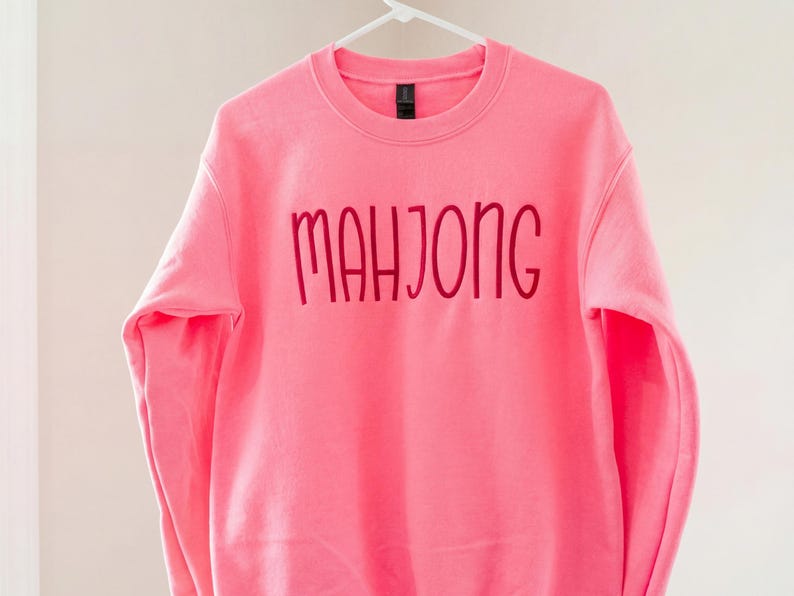 May include: A vibrant pink sweatshirt featuring the word "MAHJONG" in a darker pink, cursive script. The garment is suspended on a white hanger, set against a simple backdrop.