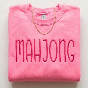 May include: A folded pink sweatshirt embroidered with the word "MAHJONG" in a darker pink. A gold chain necklace rests on the top of the sweatshirt. The sweatshirt is a solid colour and has a crew neck.