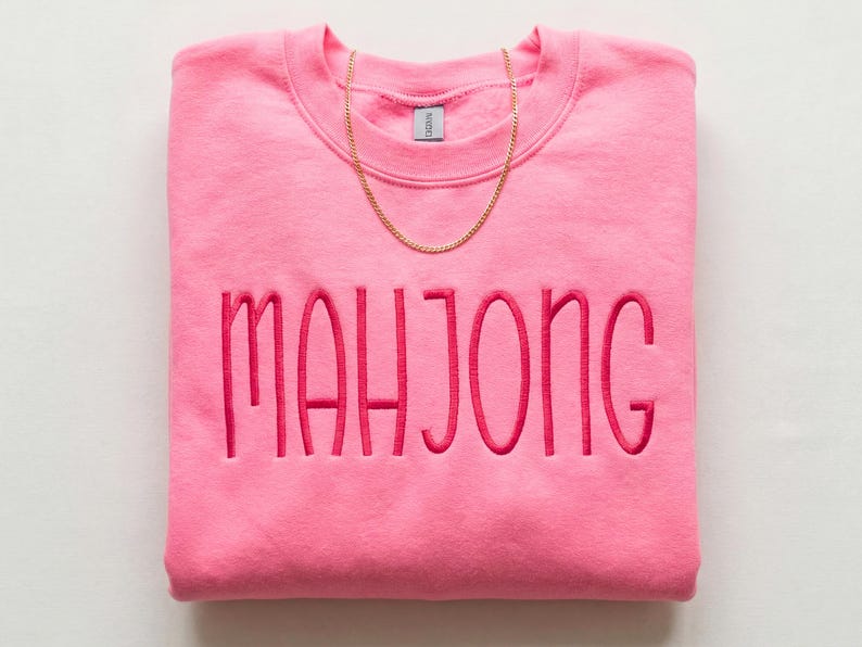 May include: A folded pink sweatshirt embroidered with the word "MAHJONG" in a darker pink. A gold chain necklace rests on the top of the sweatshirt. The sweatshirt is a solid colour and has a crew neck.