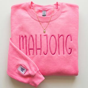 May include: A vibrant pink sweatshirt embroidered with the word "MAHJONG" in pink thread. A gold necklace with a square pendant is placed on the garment. The left sleeve has a small patch with Asian characters.