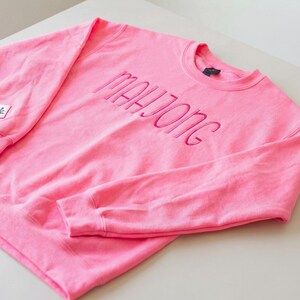 May include: A vibrant pink sweatshirt featuring the embroidered word "MAHJONG" in matching pink thread across the chest. The crew neck sweatshirt has long sleeves. A small square patch with a symbol is on the left sleeve.