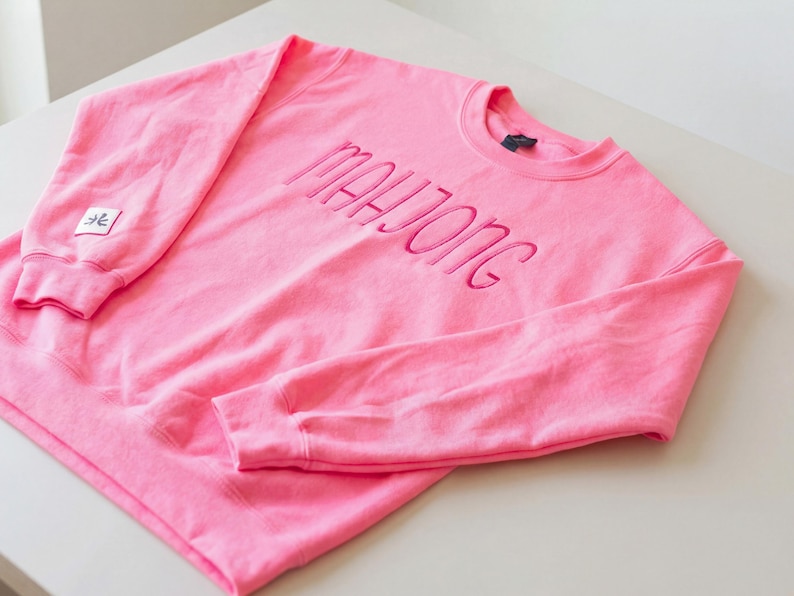May include: A vibrant pink sweatshirt featuring the embroidered word "MAHJONG" in matching pink thread across the chest. The crew neck sweatshirt has long sleeves. A small square patch with a symbol is on the left sleeve.