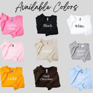 May include: Nine crewneck sweatshirts are shown in a range of colours: Safety Pink, Black, White, Light Pink, Sand, Sport Grey, Gold, Dark Chocolate, and Light Blue. The text "Available Colors" is at the top.