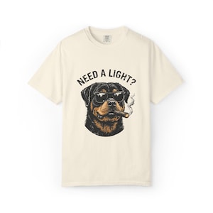 May include: Cream-colored t-shirt featuring a graphic of a Rottweiler wearing sunglasses and smoking a cigar. The text "NEED A LIGHT?" is above the dog's head in black letters. The shirt has a crew neck and short sleeves.